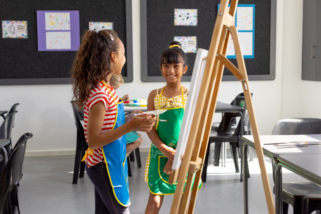 In school, in art class, biracial and a Caucasian girl, both young, are painting. Both wearing aprons, biracial girl with dark curly hair holds a palette, unaltered